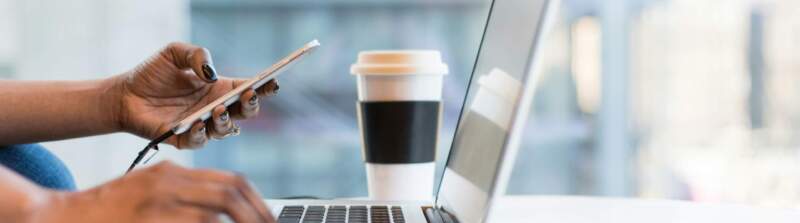 Person arbeitet am Laptop und hält Smartphone, Kaffeebecher und Brille liegen auf dem Tisch in heller Büroumgebung.