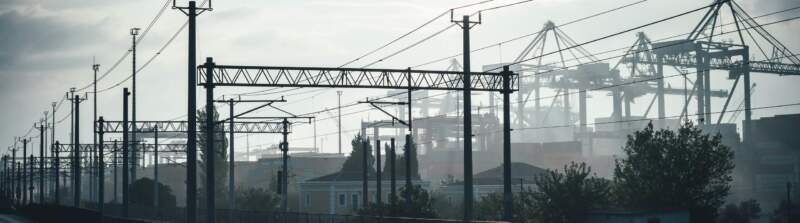 Bahngleise mit Oberleitungen im Vordergrund, Containerkräne und Hafenanlagen im Hintergrund bei Nebel und Gegenlicht.