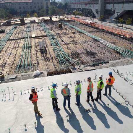 Bauarbeiter in Warnwesten und Helmen stehen auf einer großen Baustelle mit Bewehrung und Rohrleitungen, Diskussion und Planung von Infrastrukturarbeiten.