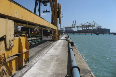 Gelbe Kranbahnführung am Kai mit Wasser und Containerbrücken im Hintergrund, Hafenlogistik bei klarem blauen Himmel.