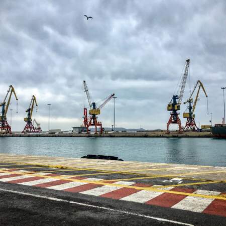 Industriehafen mit mehreren großen Kränen und Frachtschiffen an einem Kai bei bewölktem Himmel, Blick über das Wasser auf die Hafenanlagen.