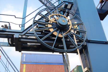 Große Kabeltrommel an einem blauen Hafenkran, im Hintergrund bunte Containerstapel auf dem Terminal.