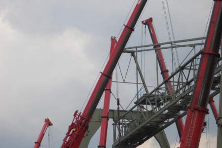 Mehrere rote Mobilkrane heben eine große Stahlkonstruktion in die Höhe vor grauem Himmel, Baustelle für schwere Montagen.