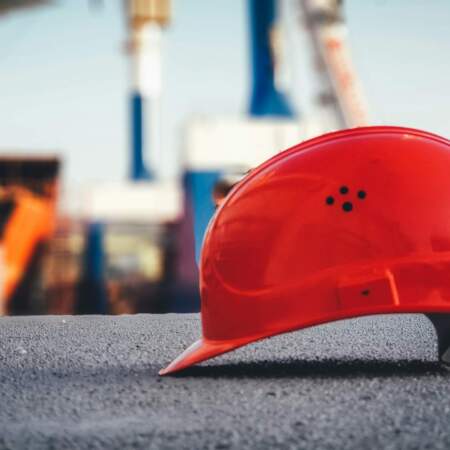 Roter Schutzhelm auf Asphalt im Vordergrund, unscharfer Hintergrund mit Kränen und Industrieanlage.