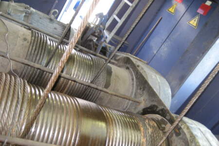 Detailaufnahme von Stahl-Drahtseilen auf massiven Seiltrommeln in einer blauen Industrieanlage mit Warnschildern.