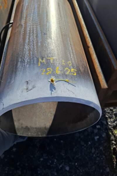 Nahaufnahme eines Stahlrohrs mit gelber Beschriftung und Datum, daneben eine kleine Blume, Industrieumgebung im Freien.