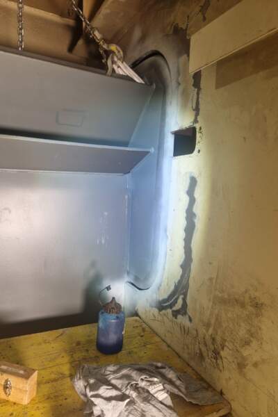 Innenraum mit geschweißtem und angeschliffenem Bereich an einer Stahlwand, Kette mit Haken, Werkbank mit Tuch und Lampe.